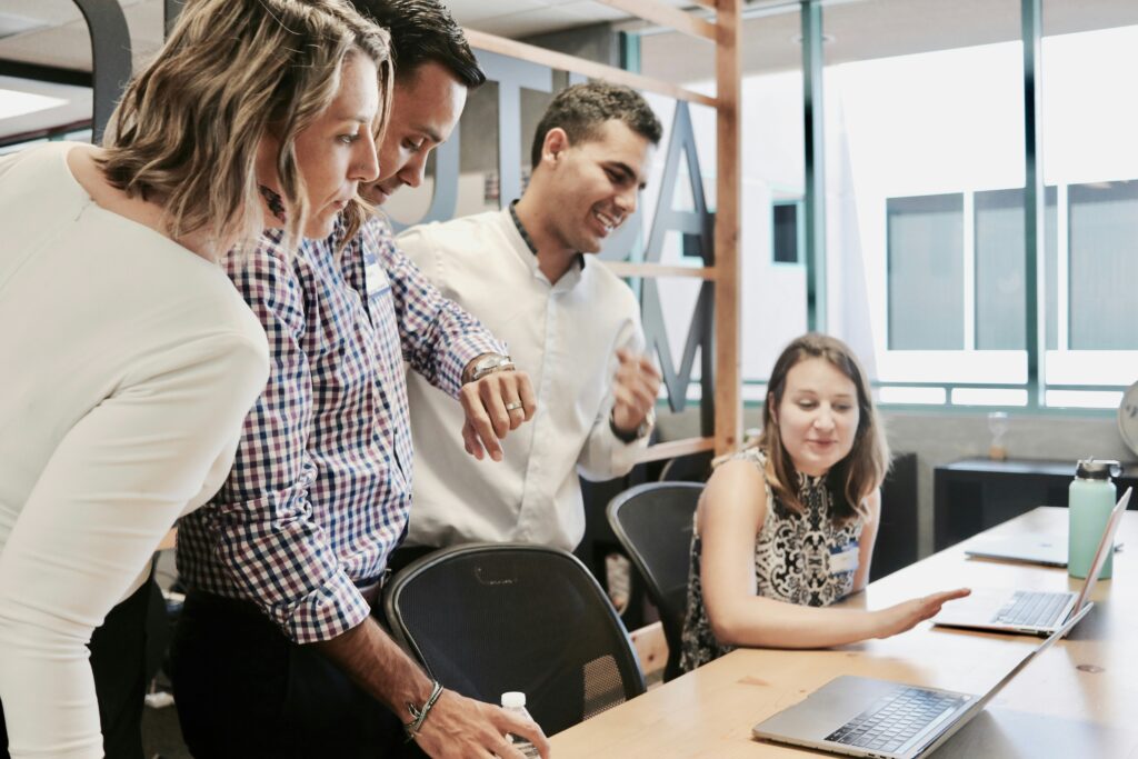 A photograph of two woman and two men at a desk looking at an unknown item on a shared computer.