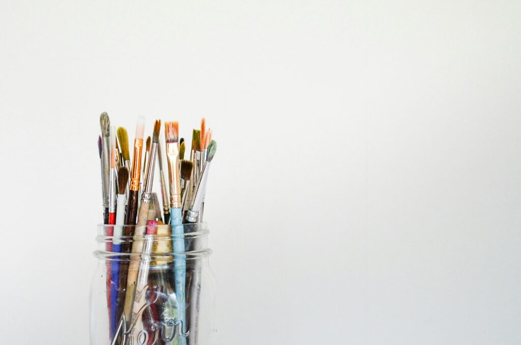 A photograph of a glass jar holding many different paint brushes, and a white wall background,